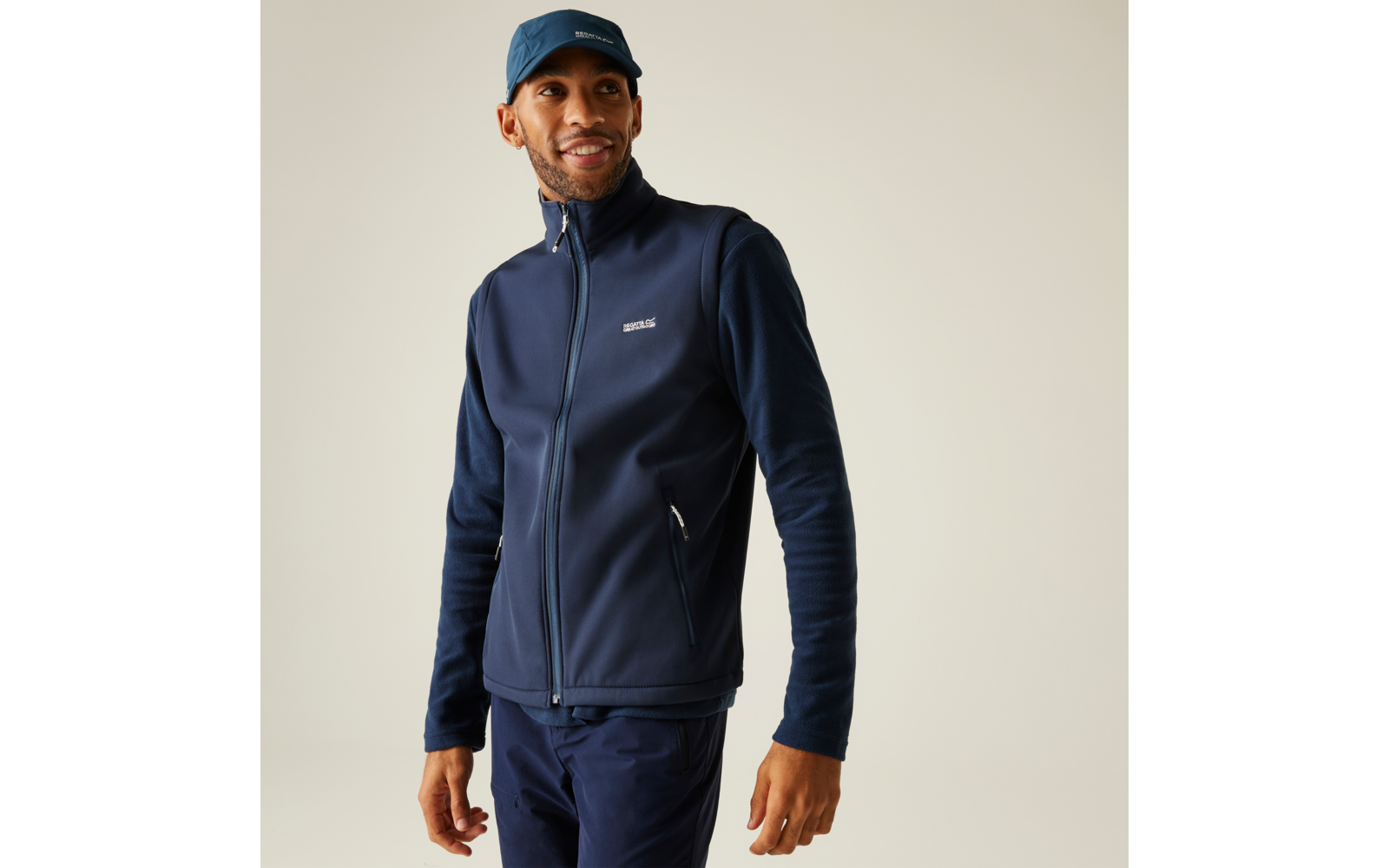 Mann vor hellem Hintergrund, trägt dunkelblaue Zip‑Jacke mit langen Ärmeln, zwei Reißverschlusstaschen und kleinem Brustlogo, dunkelblaue Kappe und Hose, blickt nach rechts