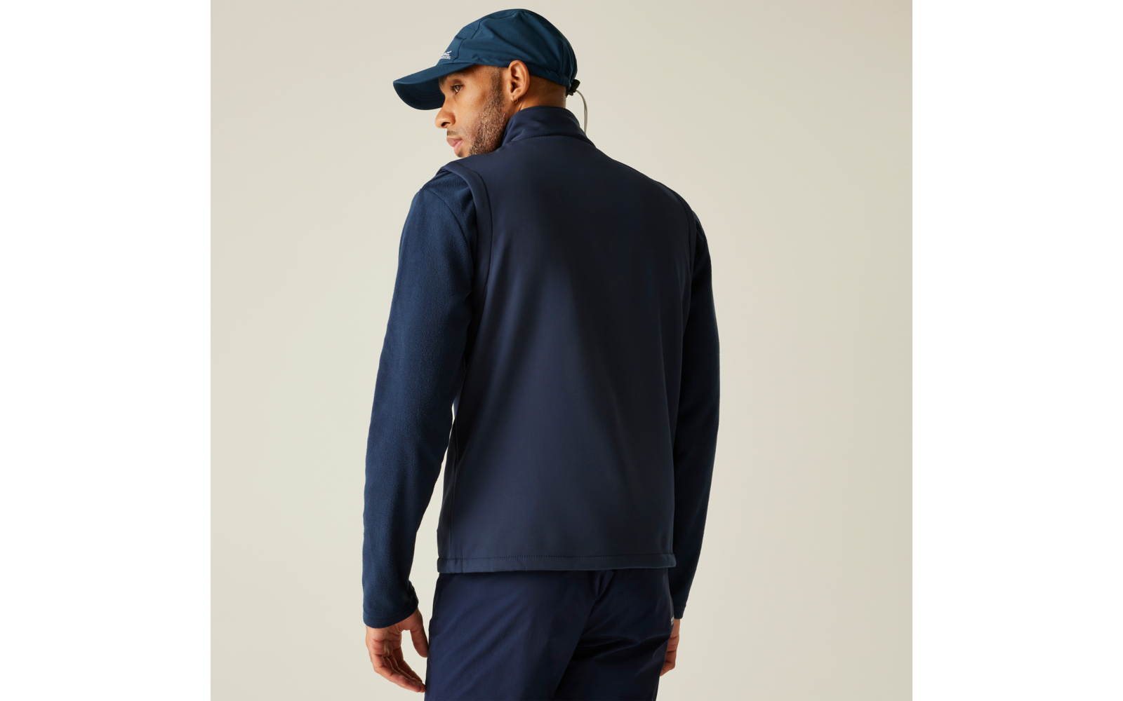 Person von hinten trägt dunkelblaue Weste über langärmeligem Shirt, dunkelblaue Hose und Baseballcap, heller Hintergrund