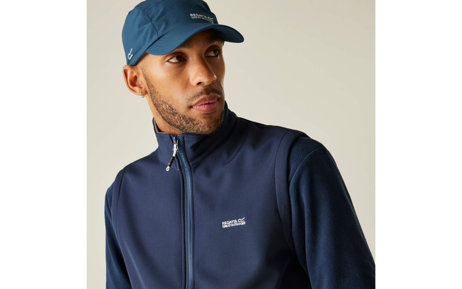 Mann mit kurzem Bart trägt dunkelblaues Cap und Reißverschlussjacke mit sichtbarem Logo, schaut nach rechts vor hellem Hintergrund