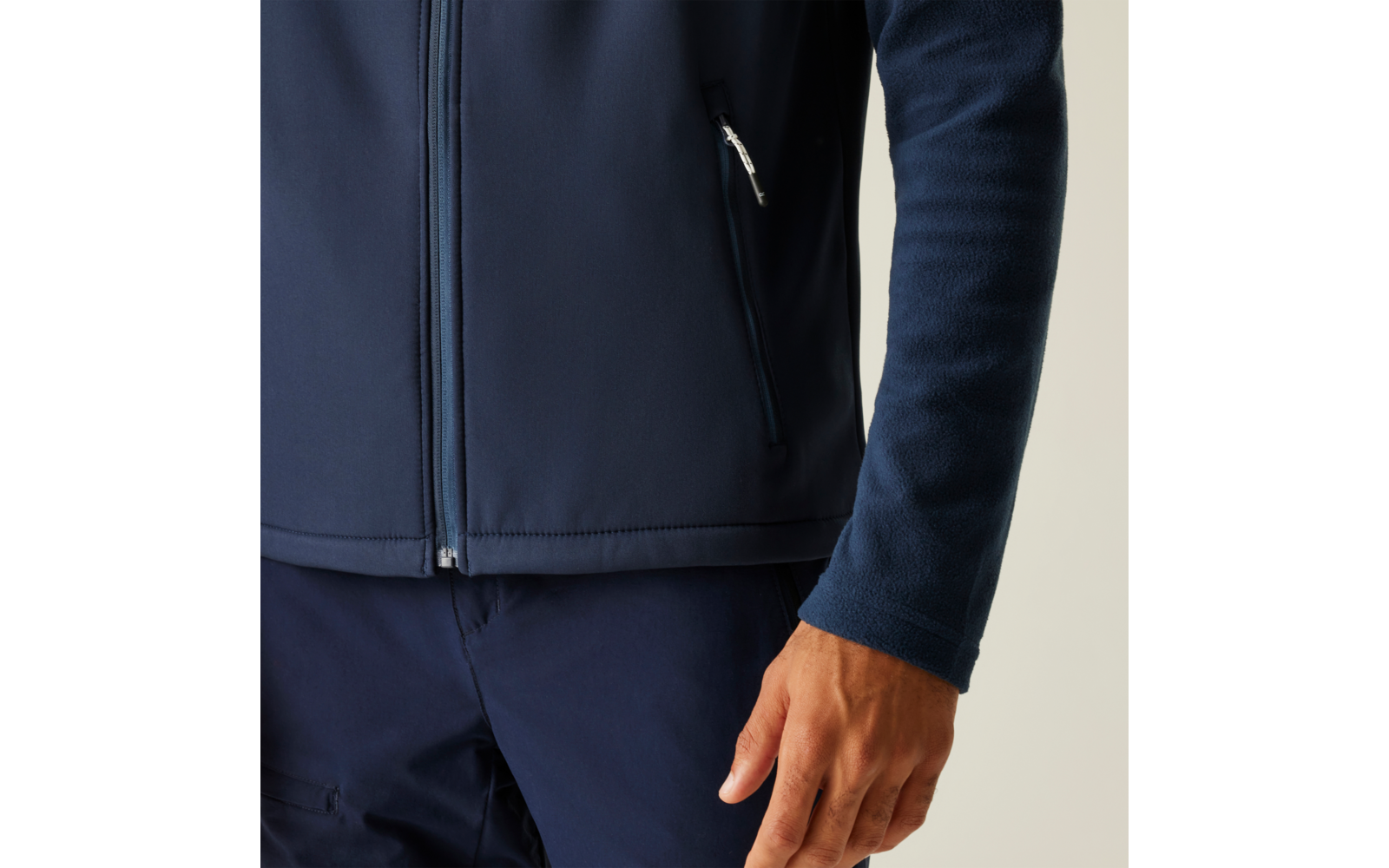 Oberkörper und rechter Arm einer Person mit dunkelblauer Jacke mit Reißverschluss und dunkler Hose, sichtbare Hand, einfarbiger heller Hintergrund