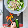 Vogelperspektive: Hände schneiden eine Gurke auf einem Schneidebrett; daneben geschnittene rote und gelbe Cherrytomaten. Oben eine Plastikschüssel mit gemischtem Salat, Gurkenscheiben, roten Zwiebelringen und hellen Bohnen