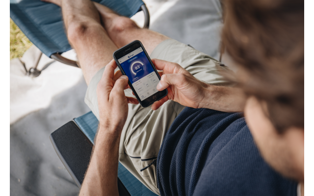 Person sitzt auf einer Liege und hält ein Smartphone mit sichtbarem blauem App‑Bildschirm und rundem Anzeigeinstrument, trägt dunkelblaues Shirt und beige Shorts; schwarzes Tablet auf der Armlehne sichtbar