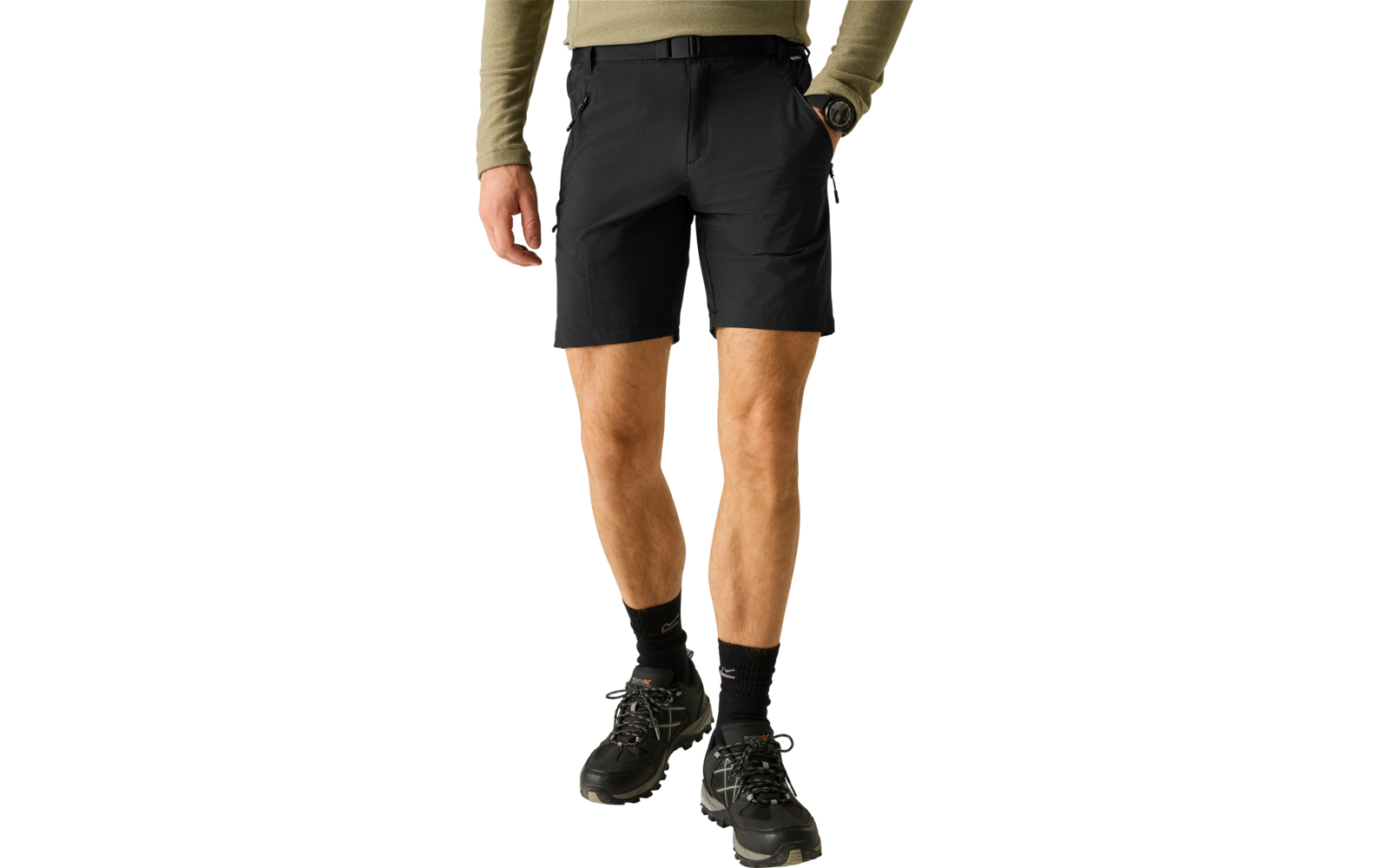 Unterkörper einer stehenden Person mit olivgrünem Langarmshirt, schwarzen Shorts mit schwarzem Gürtel, einer schwarzen Armbanduhr, schwarzen Socken und schwarzen Wanderschuhen