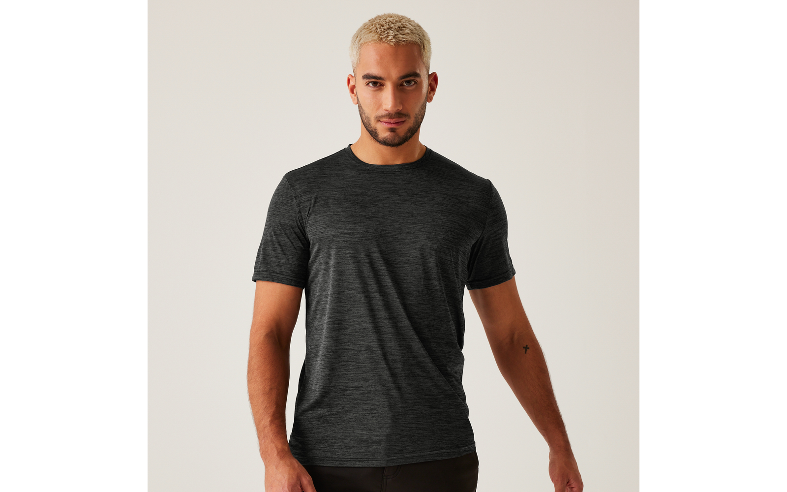 Mann mit kurz blondiertem Haar und kurzem Bart, trägt dunkelgraues meliertes T‑Shirt, steht vor hellem Hintergrund, Arme seitlich, kleines schwarzes Tattoo am inneren rechten Unterarm