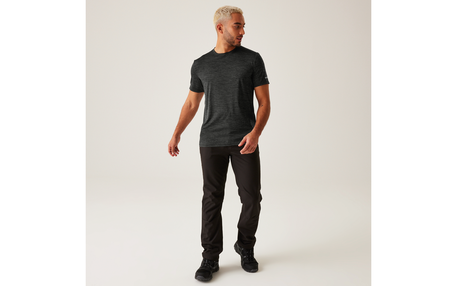 Person mit kurzem blondem Haar, dunkelgrauem T-Shirt, schwarzer Hose und schwarzen Schuhen, geht und schaut nach links