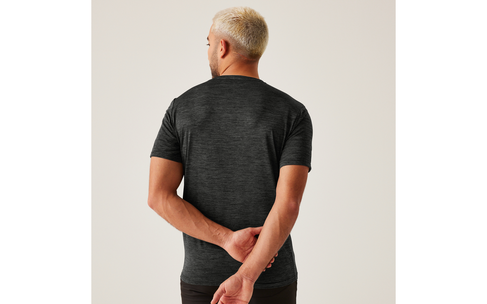 Rückansicht einer Person mit kurzgeschorenem blondem Haar, dunkelgrauem T‑Shirt mit kurzen Ärmeln, Hände hinter dem Rücken zusammengeführt, heller Hintergrund