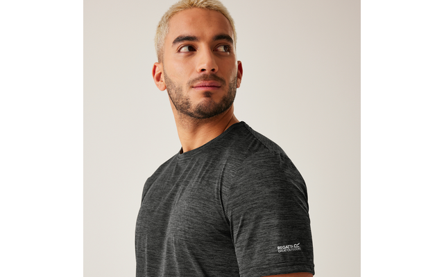 Person mit kurz blond gefärbten Haaren und kurzem Bart blickt nach links, trägt ein dunkelgraues T-Shirt mit kurzen Ärmeln, heller einfarbiger Hintergrund