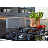 Placa de cocción de vidrio negro con tres quemadores de gas, aparato rectangular plateado perforado sobre la encimera, bolsa de papel con albahaca y tres tomates rojos, armario de vajilla al fondo