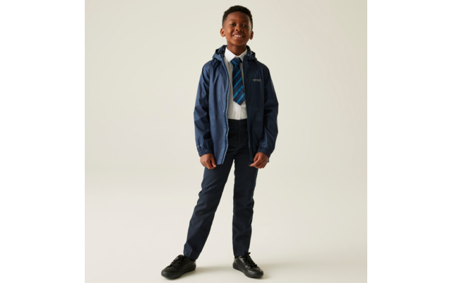 Ein Junge steht frontal mit den Händen in den Taschen, trägt eine blaue Jacke, dunkelblaue Hose und schwarze Schuhe
