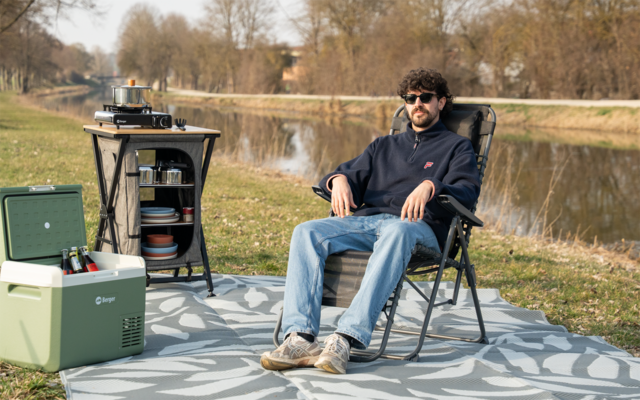 Persoon met zonnebril zit in een vouwstoel op een kleed bij een kanaal; naast hen een draagbare koelbox, een campingkooktoestel en servies op een klein tafeltje