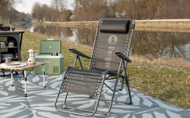 Opvouwbare donkergrijze ligstoel met hoofdkussen op een patroonmat aan de oever van een rivier; links een groene koelbox, klein tafeltje met kopjes en een lantaarn; op de achtergrond bomen en gras
