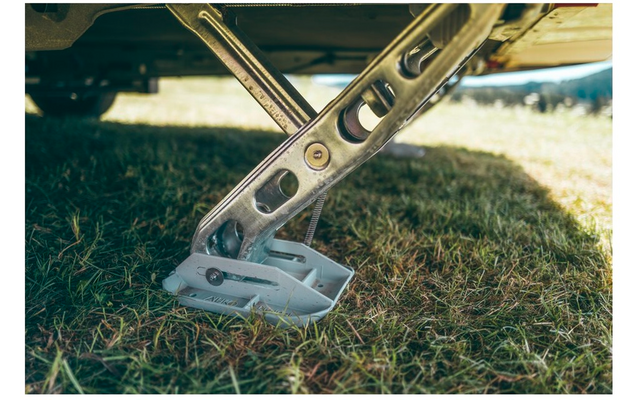 Pata de soporte metálica inclinada con piezas perforadas, fijada a una parte inferior, placa de pie de plástico blanco sobre césped, luz y sombra al fondo