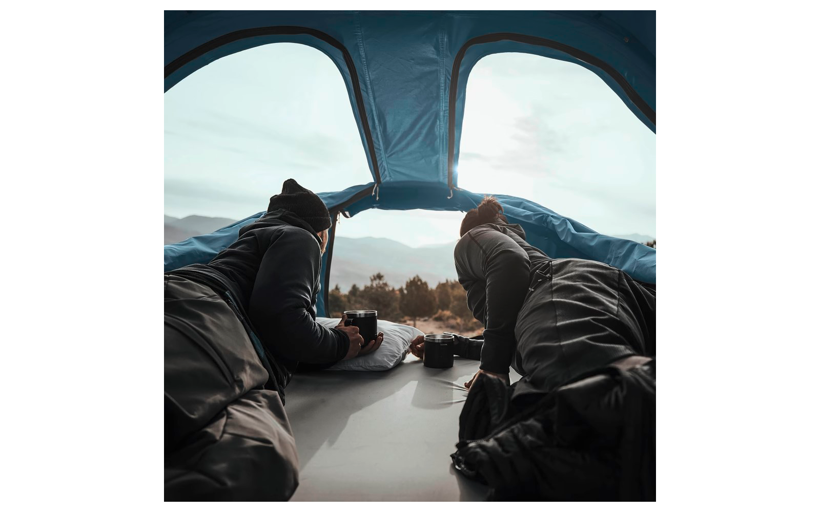 Deux personnes allongées dans une tente, visibles de dos, regardant par l'ouverture de la tente un paysage montagneux ; deux tasses et un appareil photo à côté d'eux