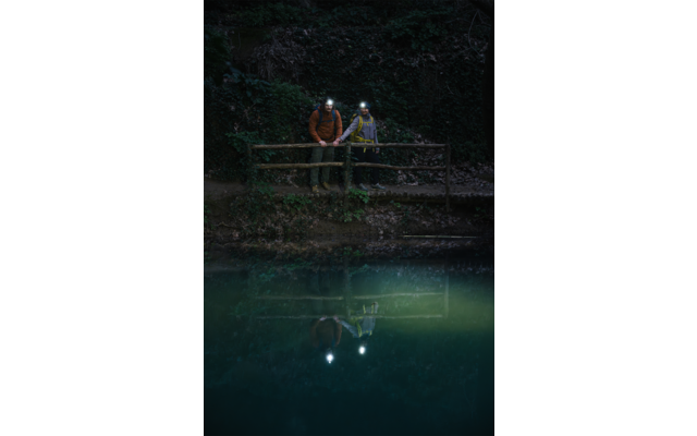 Deux personnes portant des lampes frontales allumées se tiennent près d'une rambarde en bois au bord de l'eau dans un environnement sombre ; leurs reflets et les lumières sont visibles sur l'eau, avec de la végétation derrière