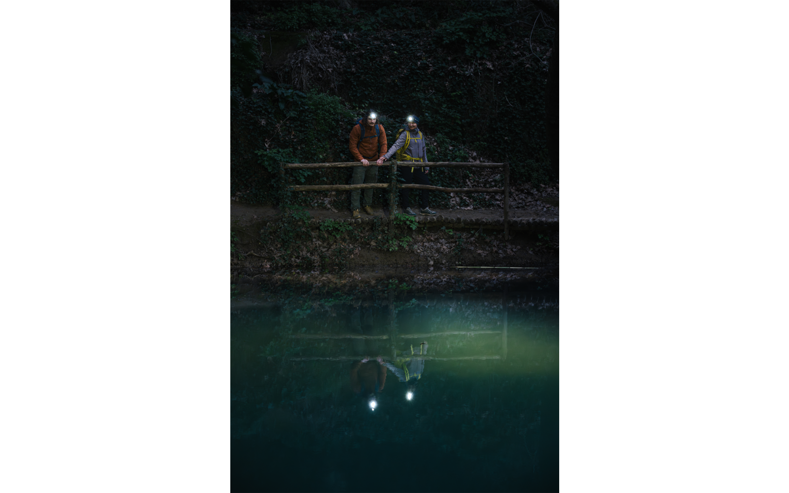 Deux personnes portant des lampes frontales allumées se tiennent près d'une rambarde en bois au bord de l'eau dans un environnement sombre ; leurs reflets et les lumières sont visibles sur l'eau, avec de la végétation derrière