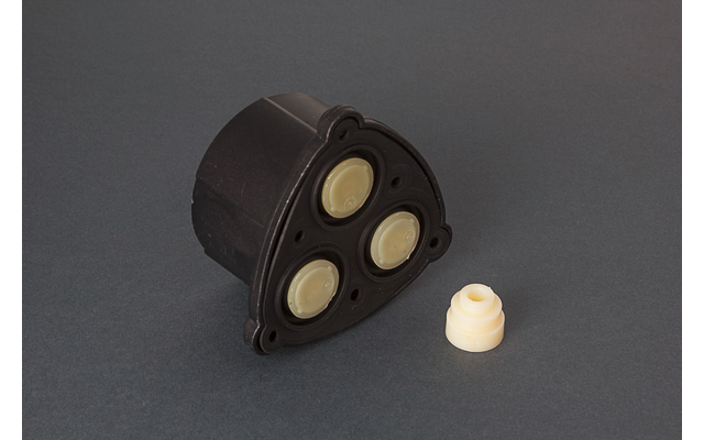 Schwarzes dreieckiges Kunststoffgehäuse mit drei beigen runden Scheiben; daneben ein kleines beiges rundes Teil auf grauem Hintergrund