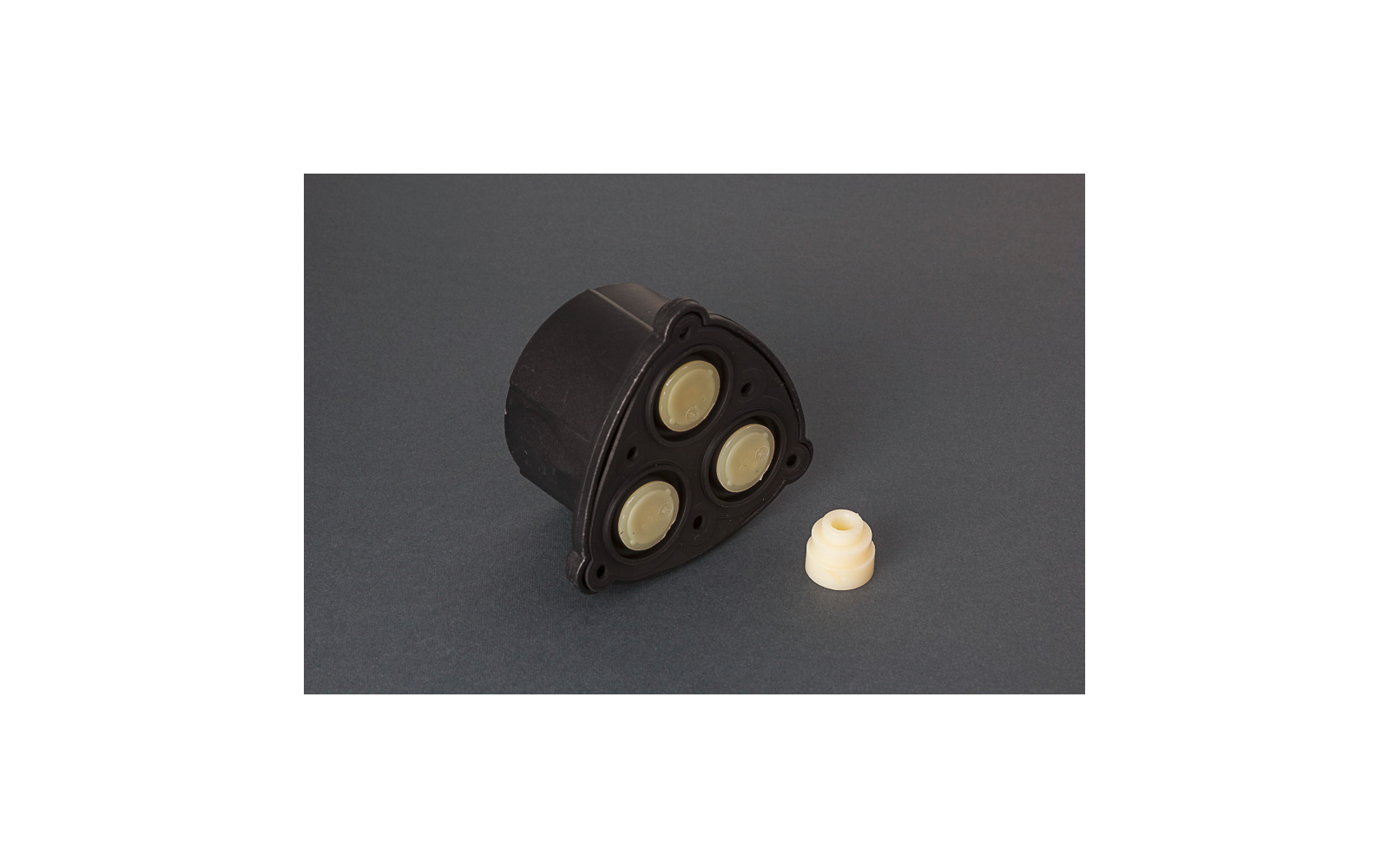Schwarzes dreieckiges Kunststoffgehäuse mit drei beigen runden Scheiben; daneben ein kleines beiges rundes Teil auf grauem Hintergrund