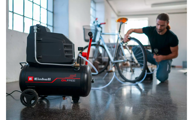 Einhell-Kompressor auf Rädern im Vordergrund; im Hintergrund kniet ein Mann und hält einen Schlauch an das Ventil eines Fahrrads