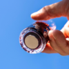 Hand hält kleines rundes Glasfläschchen mit rundem Metallplättchen an der Unterseite, blauer Himmel im Hintergrund