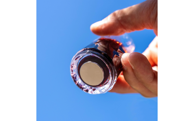 Hand hält kleines rundes Glasfläschchen mit rundem Metallplättchen an der Unterseite, blauer Himmel im Hintergrund