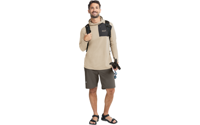 Stehender Mann, beiger Kapuzenpullover mit schwarzer Brusttasche, dunkelgraue Shorts, schwarzer Rucksack, schwarze Sandalen, hält in der rechten Hand ein schwarzes Objekt mit blauem Detail