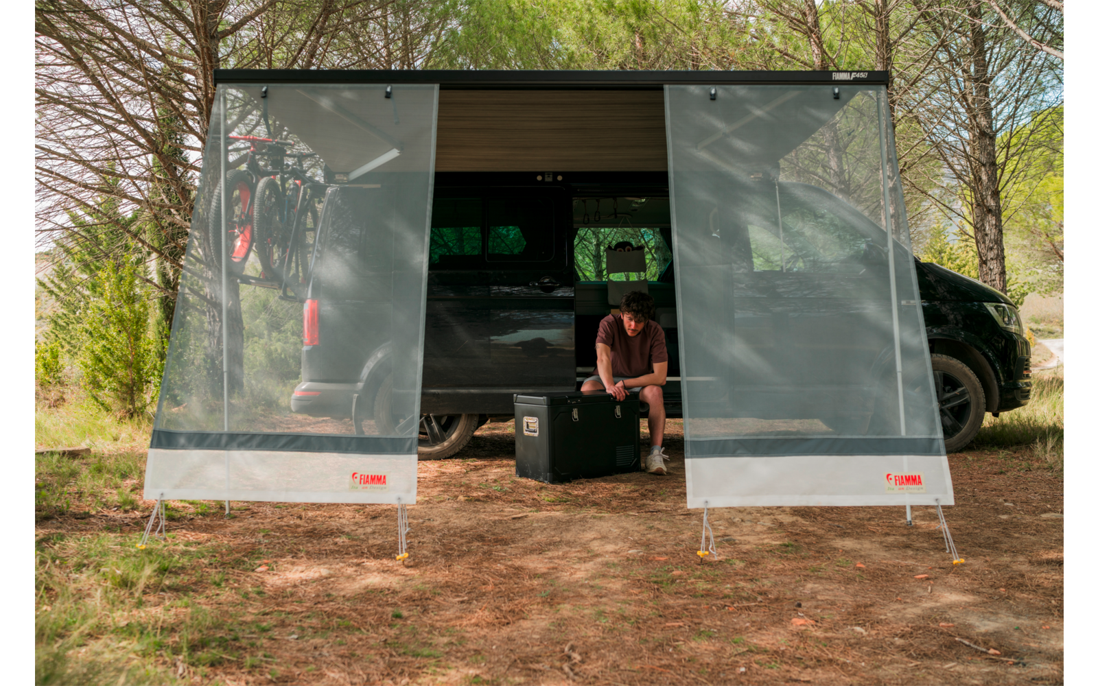 Schwarzer Van mit geöffneter Seitentür und zwei durchsichtigen Markisenpaneelen; eine Person sitzt auf einem schwarzen Koffer in der Türöffnung; Fahrrad im Van; Bäume im Hintergrund