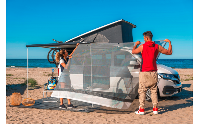 Zwei Personen befestigen ein feinmaschiges Netz an der Seite eines weißen Wohnmobils mit aufgestelltem Hochdach auf einem Sandstrand; Meer und blauer Himmel im Hintergrund; zwei Flechtkörbe im Sand