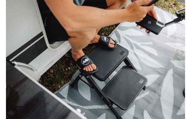 Bas des jambes et pieds d'une personne portant des sandales noires sur des marches noires, une main tient un smartphone, tapis d'extérieur gris à motifs, herbe en bordure, encadrement de porte de véhicule blanc visible