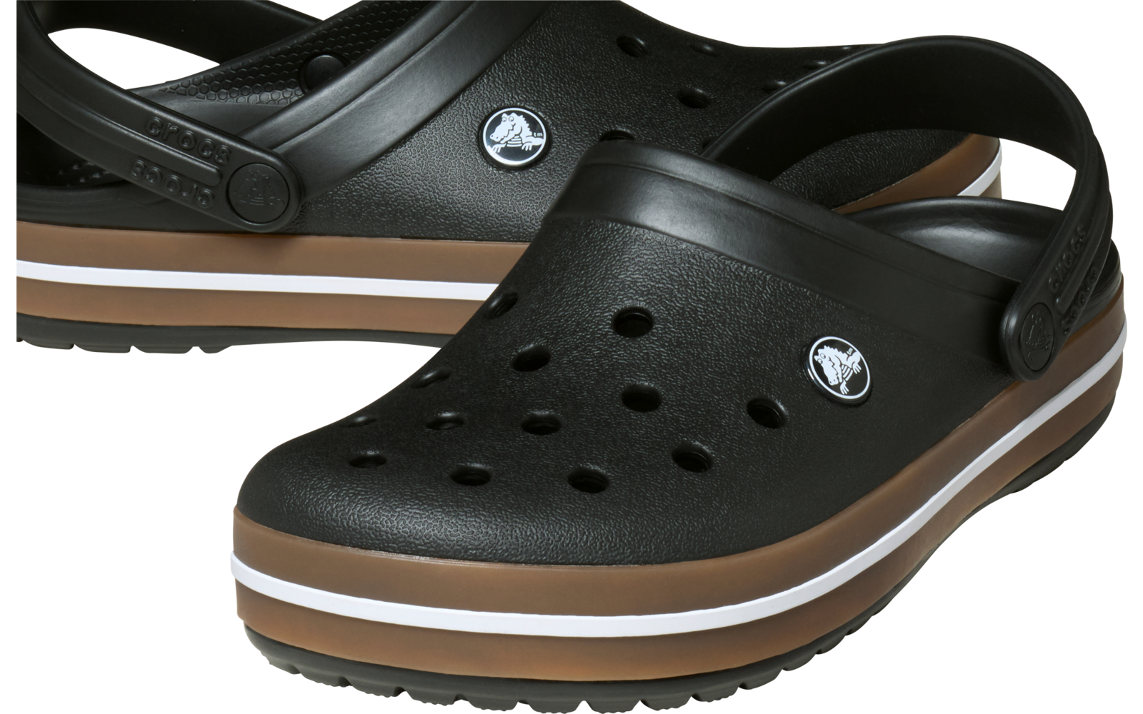 Zwei schwarze Clogs mit Fersenriemen, Löchern auf dem Vorderteil, runden Logo-Pins an den Seiten und braun-weißer Streifensohle