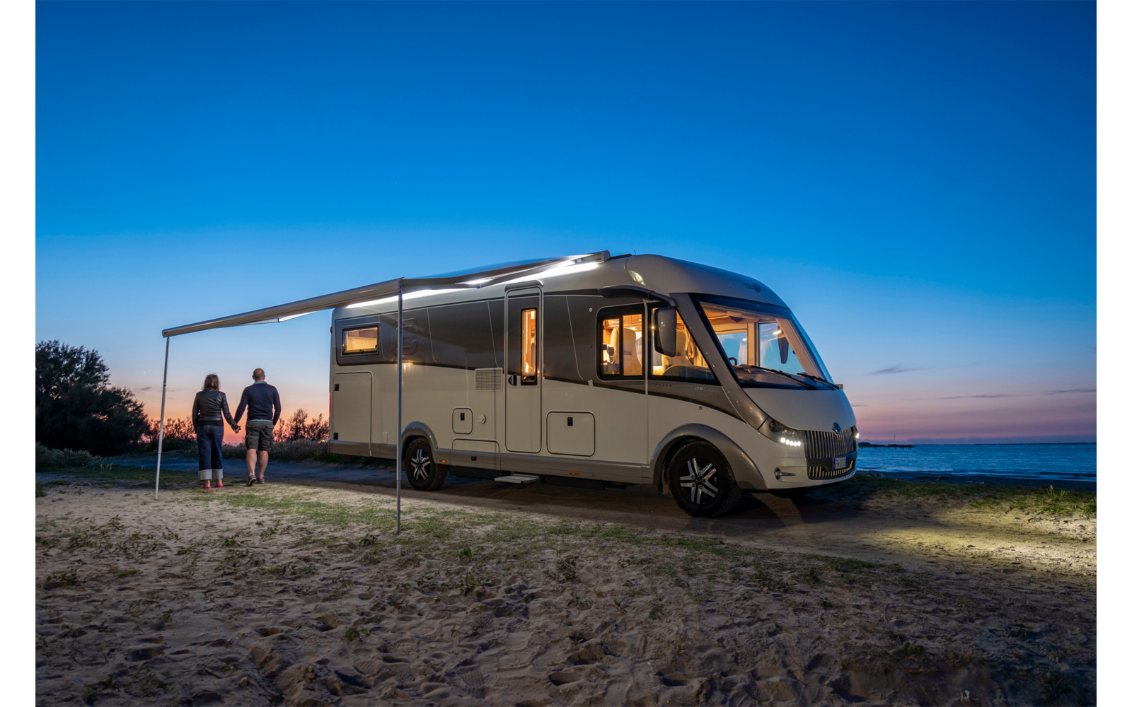 Wohnmobil steht bei Sonnenuntergang auf einem offenen Feld mit zwei Personen daneben