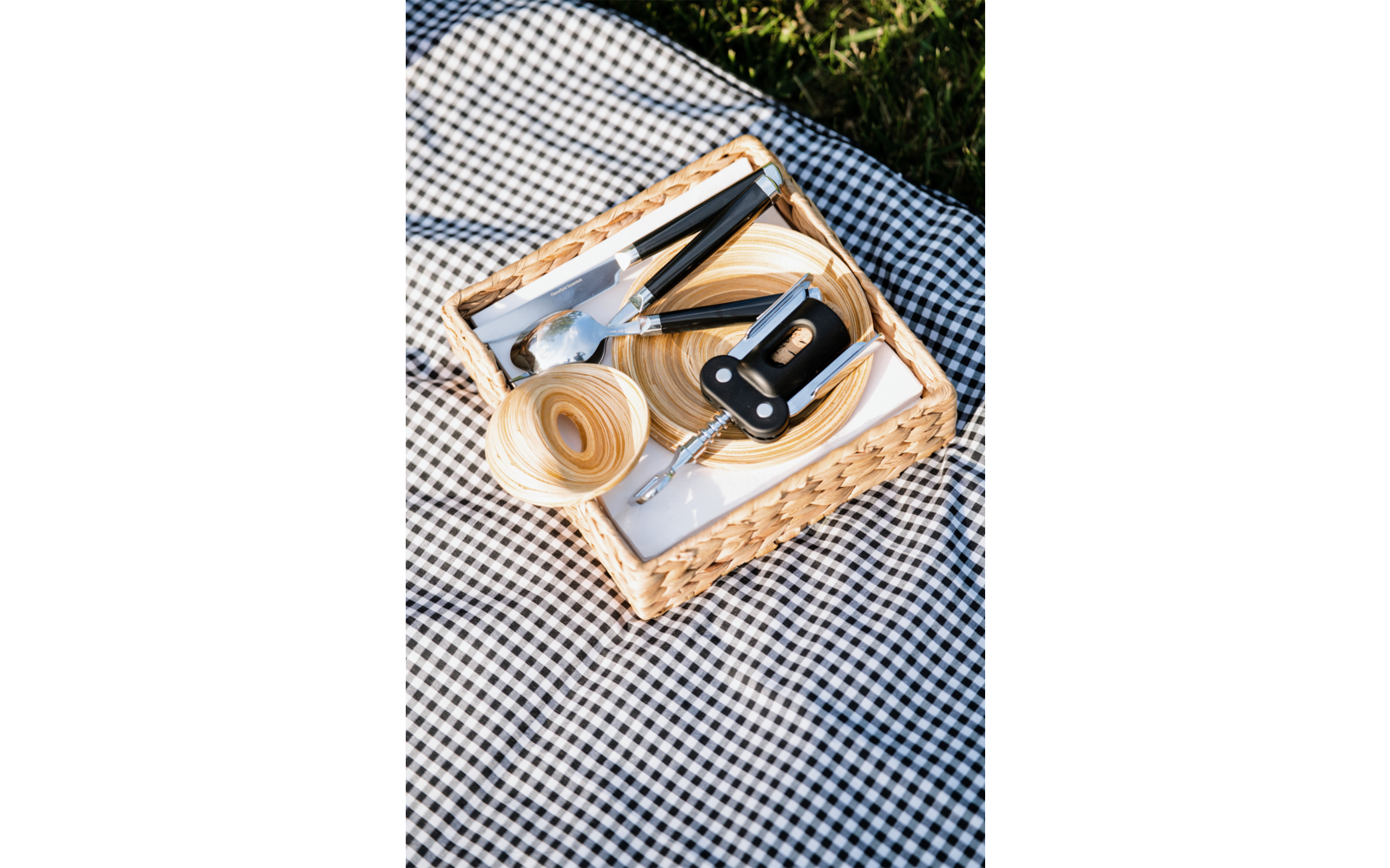 Cesto di vimini con due ciotole rotonde in legno, posate in metallo e un cavatappi nero su un panno a quadretti bianco e nero; erba sullo sfondo