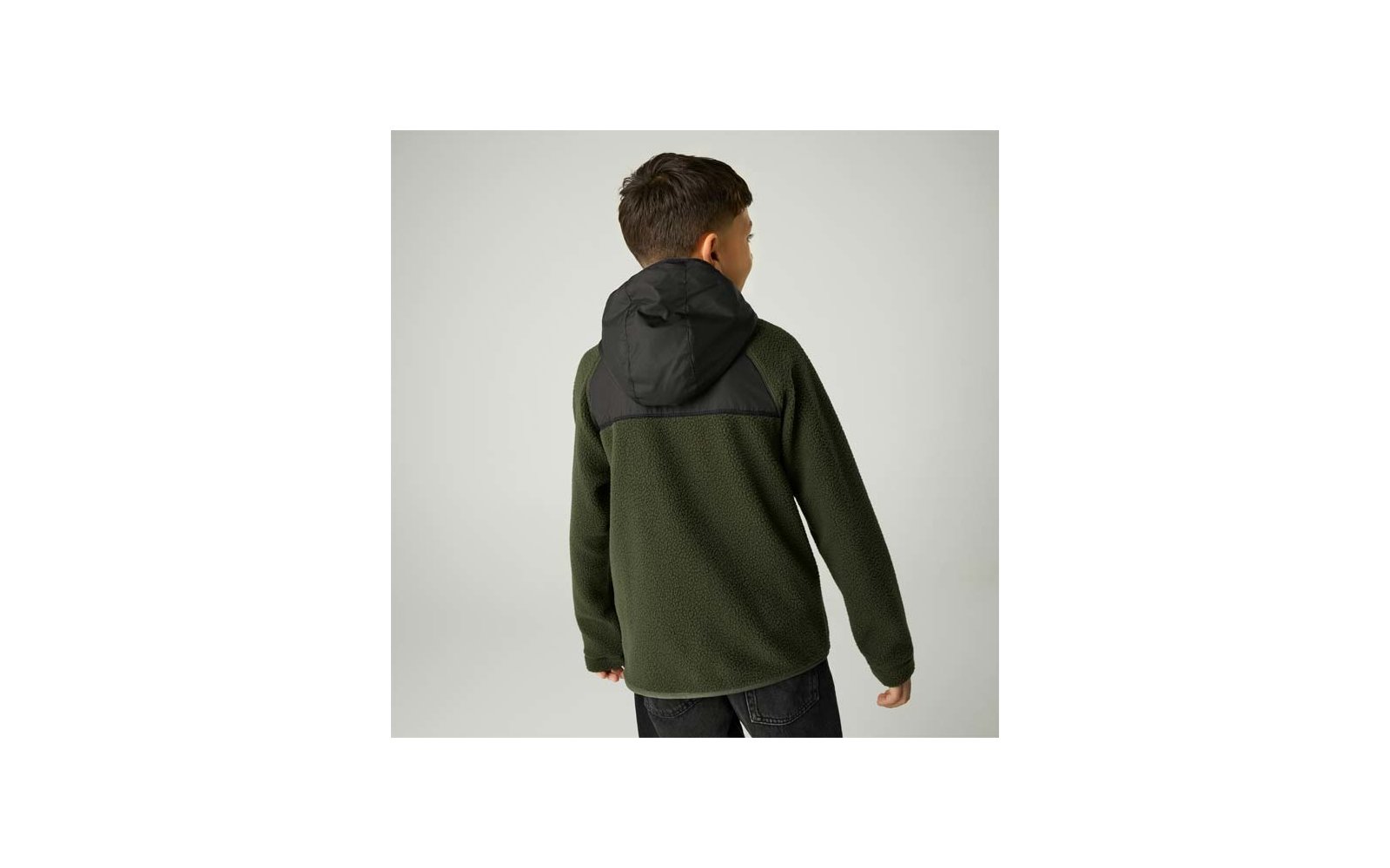 Person von hinten, trägt olivgrüne Fleecejacke mit schwarzer Kapuze und schwarzen Schultereinsätzen, vor hellem neutralem Hintergrund