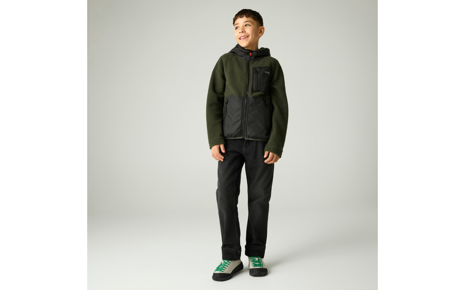 Ein Junge steht vor hellem Hintergrund, trägt eine grün-schwarze Kapuzenjacke, schwarze Hose und weiße Sneaker mit grünen Details, er lächelt