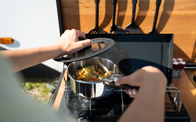 Zwei Hände heben den Deckel eines Edelstahltopfes mit Gemüse auf einem Camping-Gaskocher; im Hintergrund hängende Kochutensilien, Holzverkleidung und kleine Vorratsgläser