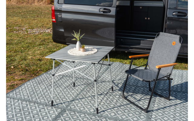 Grauer Stuhl mit Holzarmlehnen und faltbarer Tisch mit runder Matte und kleiner grüner Pflanze neben geöffneter dunkler Fahrzeugtür auf gemustertem Outdoor-Teppich; Gras im Hintergrund