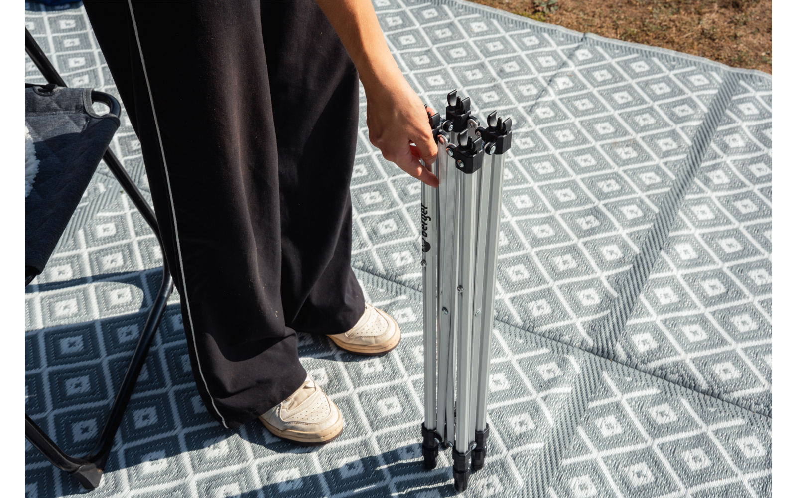 Unterkörper einer Person in schwarzen Hosen und weißen Schuhen, stehend auf einer grau-weißen, gemusterten Matte, hält ein zusammengeklapptes silbernes Stativ; Stuhlbein links, Gras oben rechts