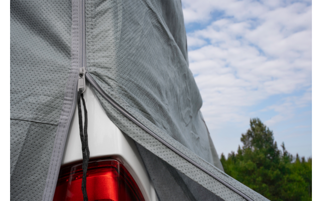 Partially pulled-back gray car cover revealing a red taillight and silver vehicle body, zipper and strap visible, cloudy sky and trees in the background