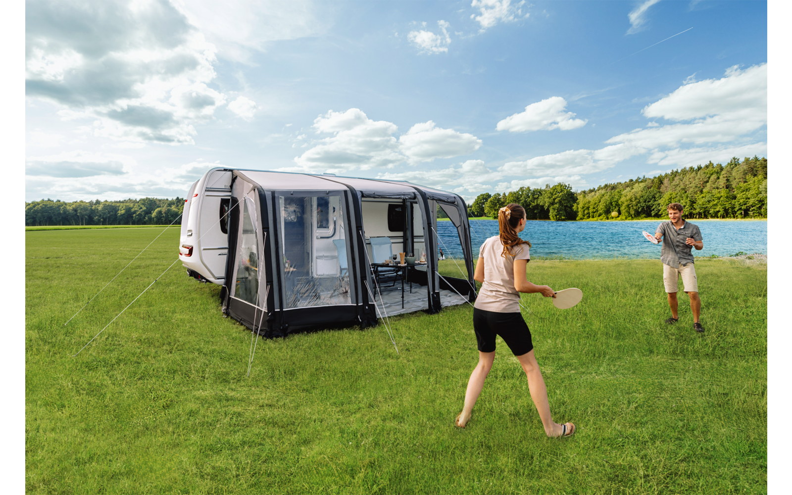 Zwei Personen auf einer grünen Wiese, jeweils mit einem Tischtennispaddel und einem Ball, neben einem Wohnwagen mit angebautem Vorzelt; im Hintergrund See und Wald