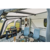 Voortent bij een caravan met twee blauwe campingstoelen, een eettafel met borden, kommen, glazen, bestek, een kom met salade, een rek met kooktoestel en voorraden, een open deur van de caravan en gras op de achtergrond