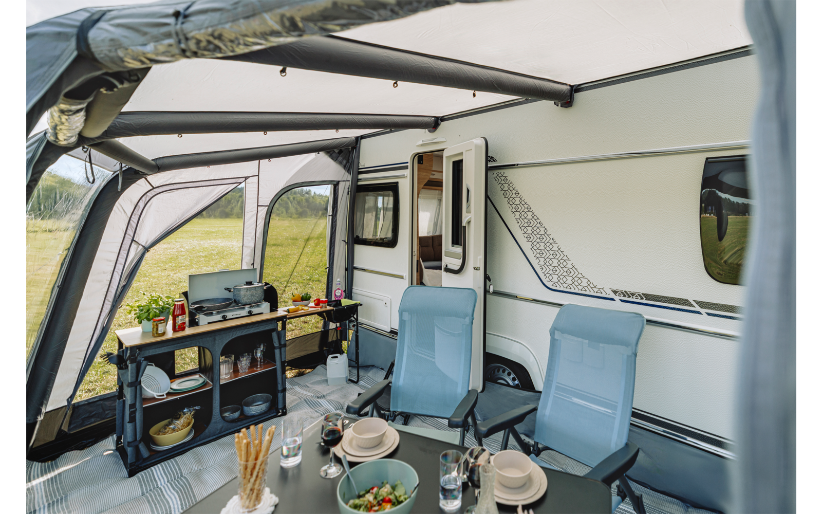 Voortent bij een caravan met twee blauwe campingstoelen, een eettafel met borden, kommen, glazen, bestek, een kom met salade, een rek met kooktoestel en voorraden, een open deur van de caravan en gras op de achtergrond