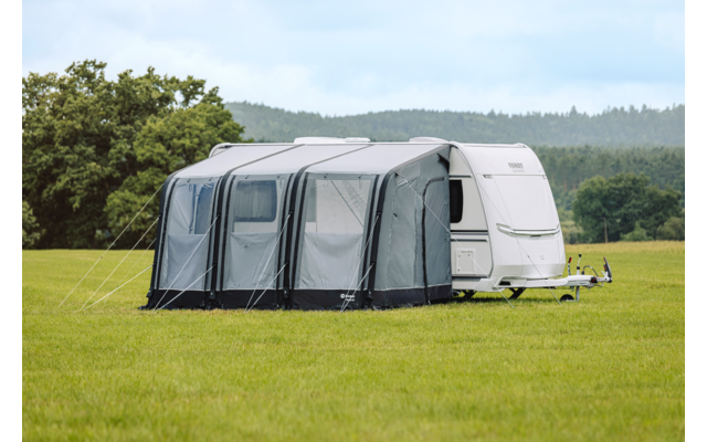 Witte caravan met een grijze voortent op een groen veld; bomen aan de horizon, bewolkte lucht