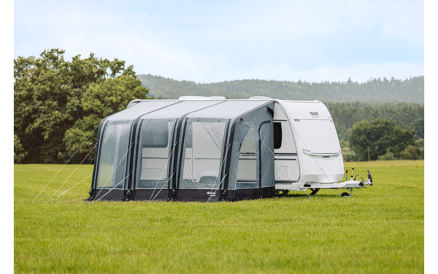 Witte caravan met grijze voortent op een groen veld, bomen en beboste heuvels op de achtergrond