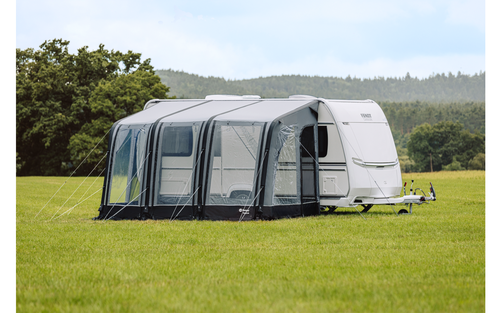 Witte caravan met grijze voortent op een groen veld, bomen en bewolkte lucht op de achtergrond