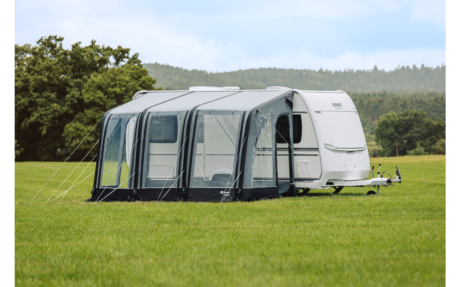 Witte caravan met grijze voortent op een groen veld, bomen en bewolkte lucht op de achtergrond