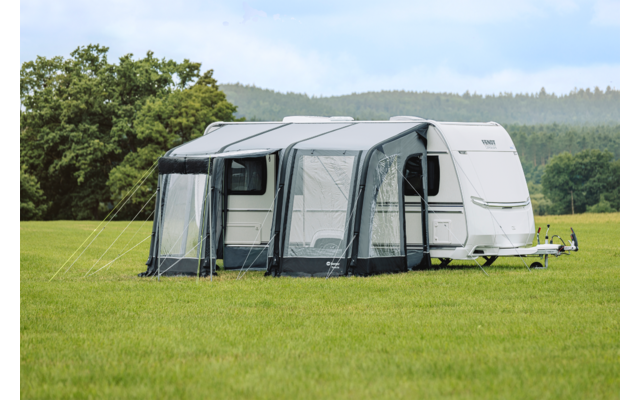 Witte caravan met een grijze voortent op een groen veld, bomen en heuvels op de achtergrond