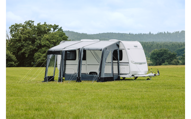 Witte caravan met een grijs voortent op een grasveld, bomen en bewolkte lucht op de achtergrond