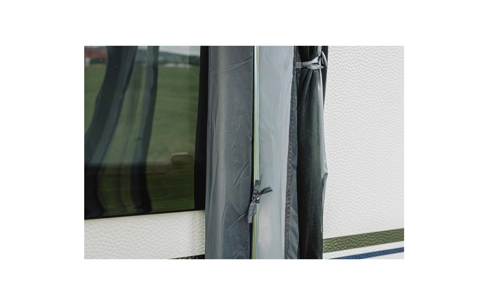 Close-up van grijs textiel met vastgeknoopt koord, een doorzichtige kunststofstrip, een witte gestructureerde wand met een blauwe streep en een donkere ruit met weerspiegeling van een groen veld