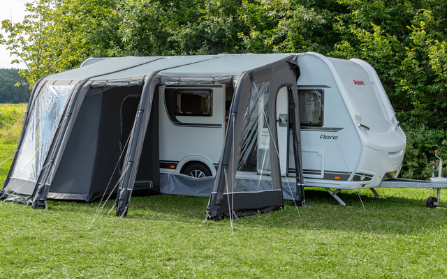 Witte caravan met een grijze voortent met grote ramen op een groen gazon voor bomen