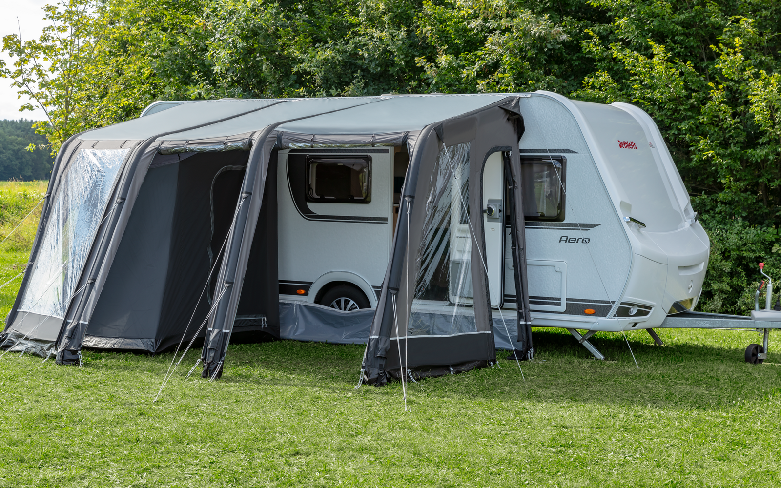 Witte caravan met een grijze voortent met grote ramen op een groen gazon voor bomen