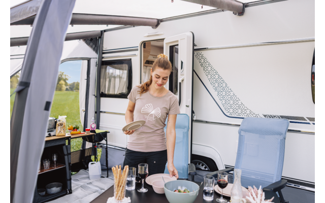 Vrouw staat bij een tafel voor een caravan met een open deur; op de tafel kommen, glazen, borden en broodstengels; opvouwbare stoel naast de tafel